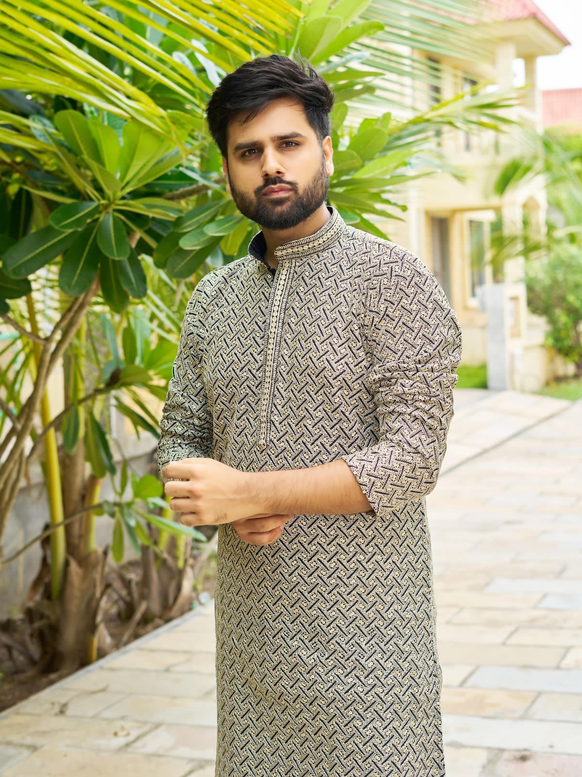 Man wearing a black patterned kurta standing outdoors with greenery in the background