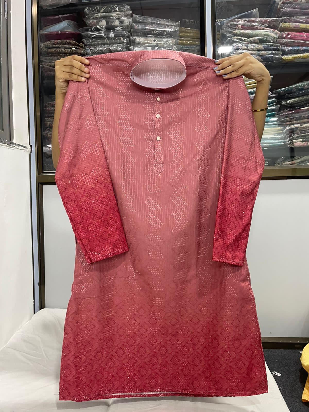 Pink kurta with intricate patterns held up against a background of folded fabric.