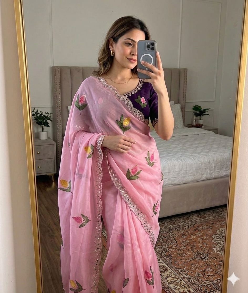 Woman taking a mirror selfie wearing a Baby pink saree with floral patterns in a bedroom.