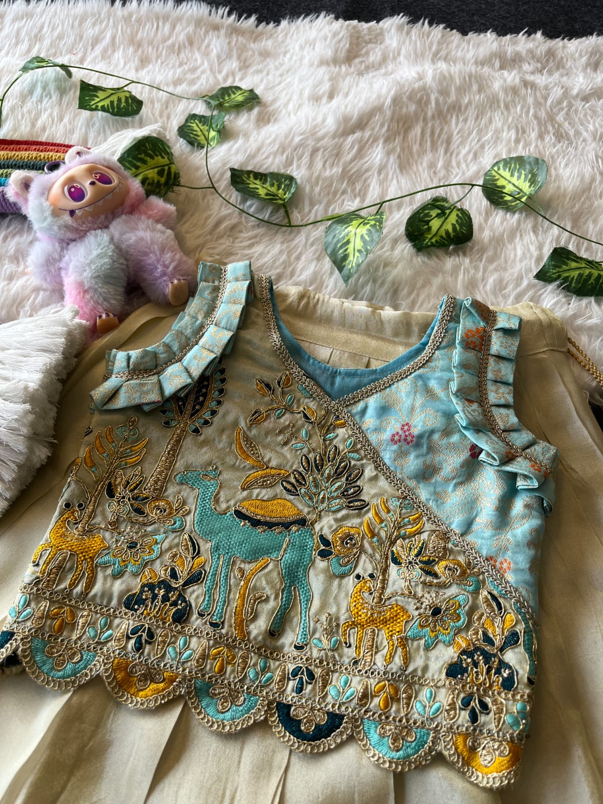 Beige Embroiderey garment with animal designs on a textured surface with a plush toy and decorative leaves.