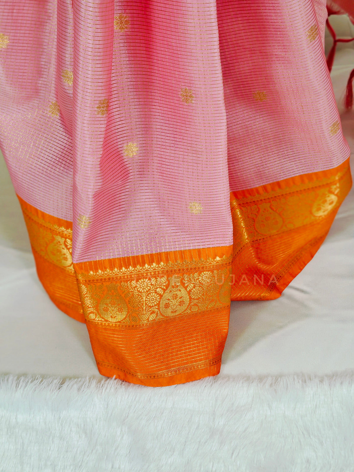A close-up of a pink saree with a wide orange border and gold zari woven motifs, draped on a white surface.