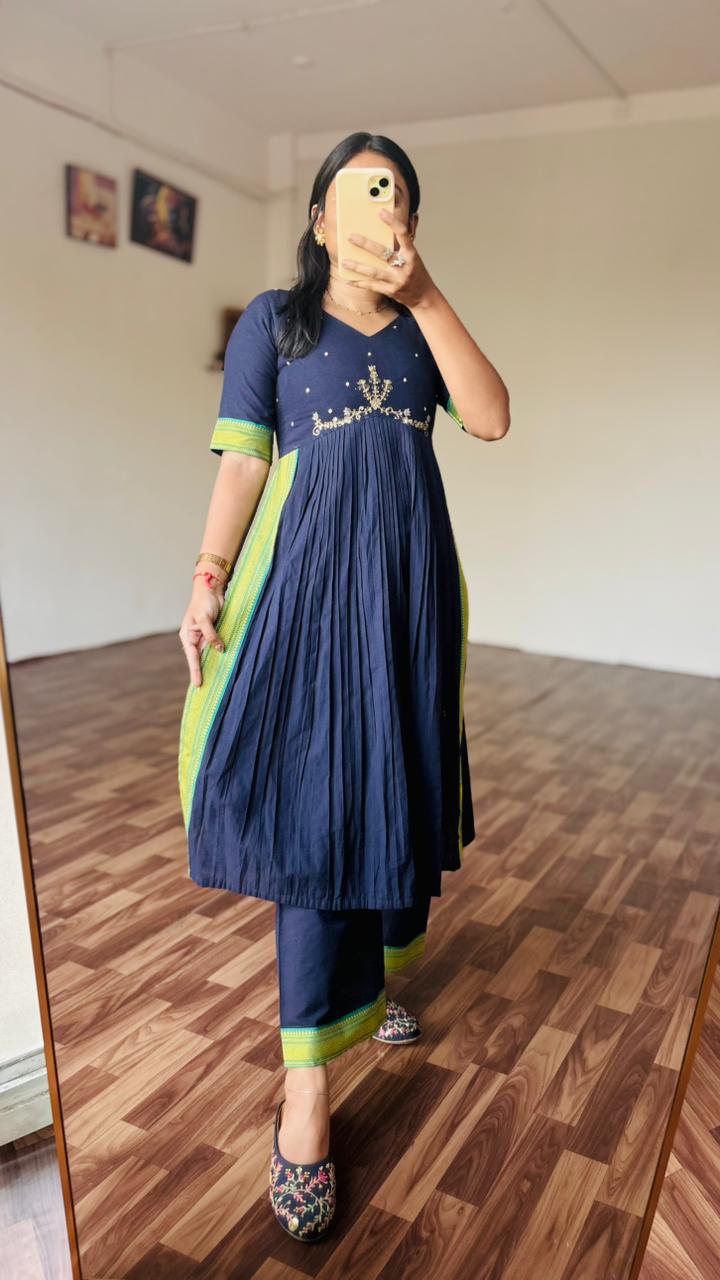 Person wearing a navy blue and green traditional outfit standing in a room with wooden flooring.