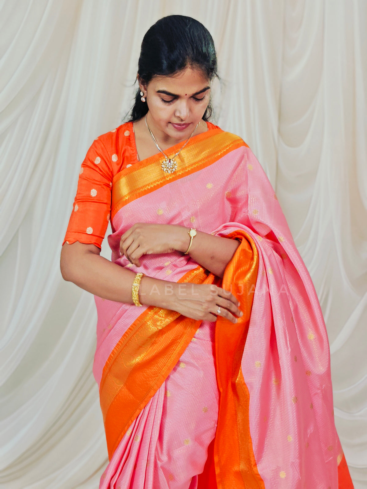 A woman wearing a pink and orange saree with gold zari detailing, standing against a draped cream fabric backdrop.