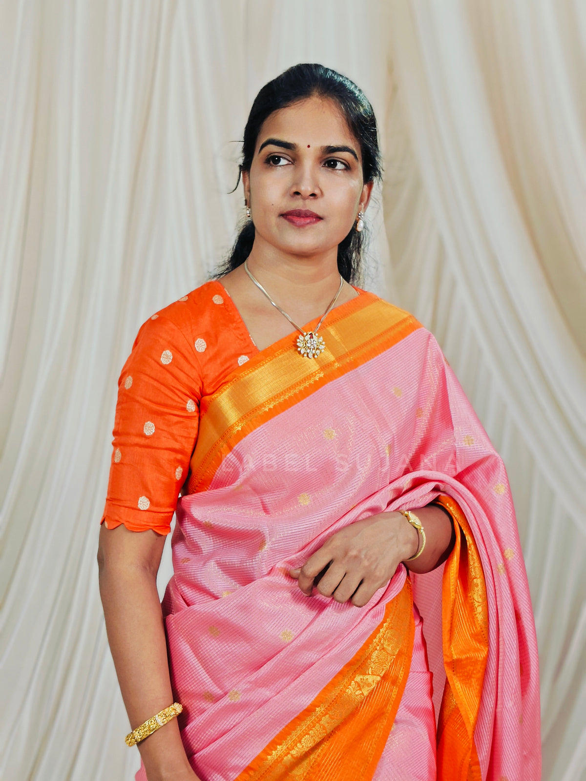 A woman wearing a pink and orange traditional sari with gold zari detailing, standing against a draped cream fabric backdrop.