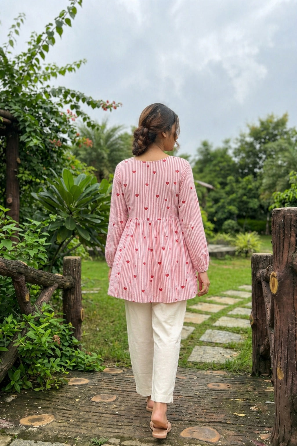 Woman in a Baby pink checkered top and white pants walking through a garden.