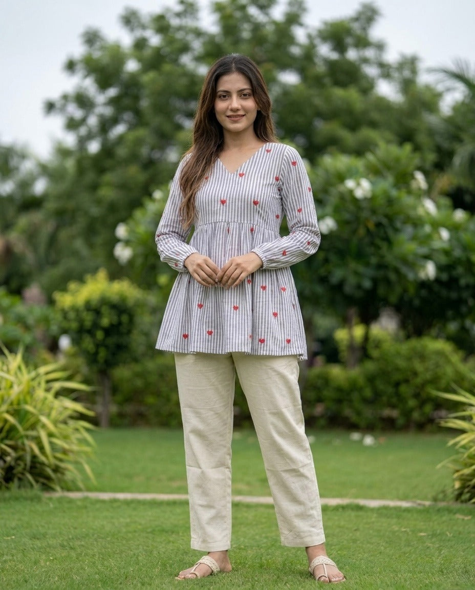 Woman standing outdoors in a garden wearing a striped gray shirt and beige pants.