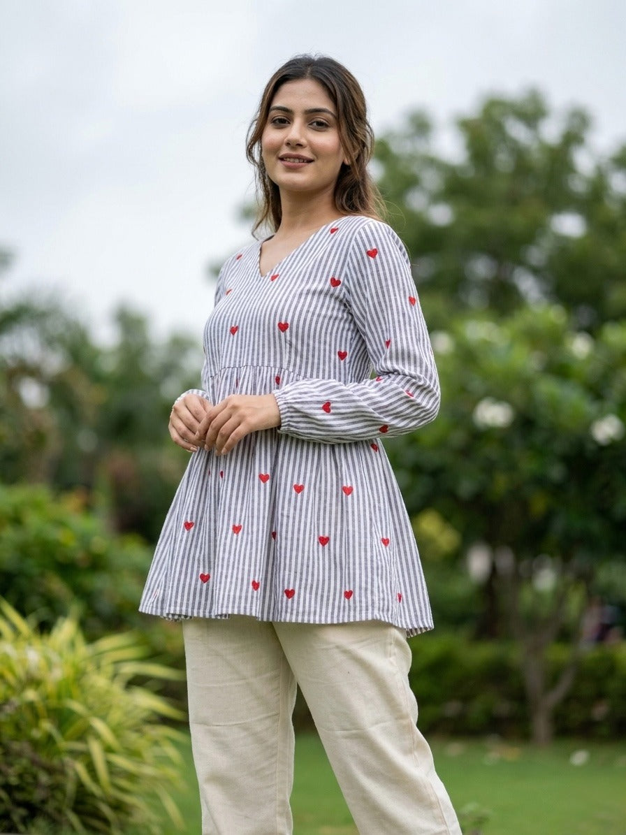 Woman wearing a gray top with red heart patterns and beige pants standing outdoors.