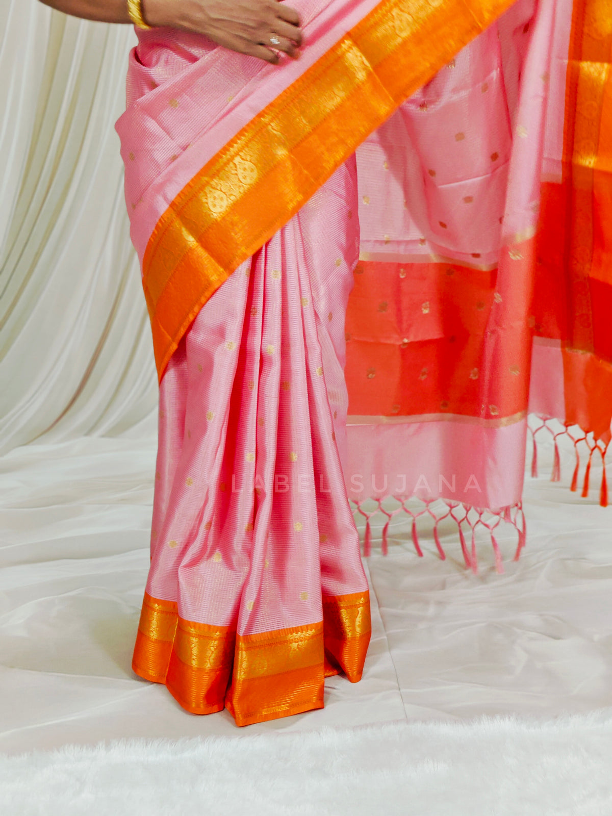 A pink silk saree with a wide orange and gold zari border, draped on a model. The fabric features small gold motifs and a matching pink and orange border with tassel ends.