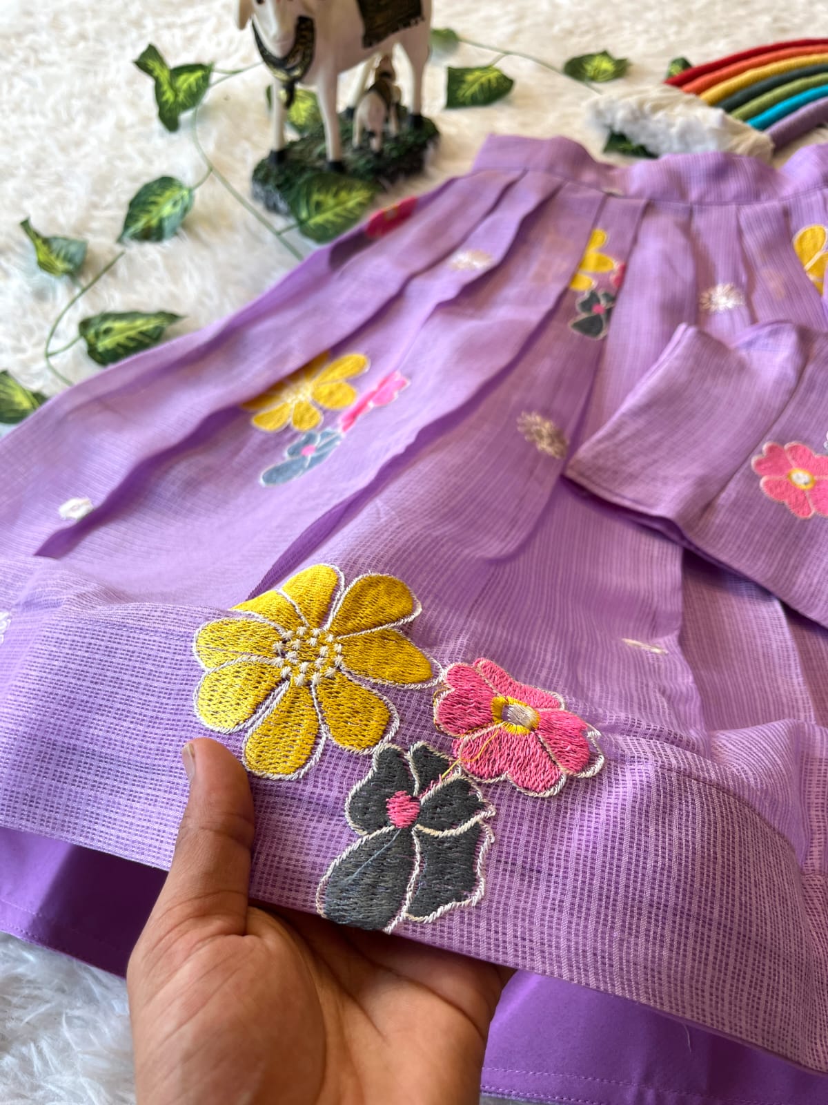 Lavender fabric with floral embroidery held by a hand, with decorative elements in the background.