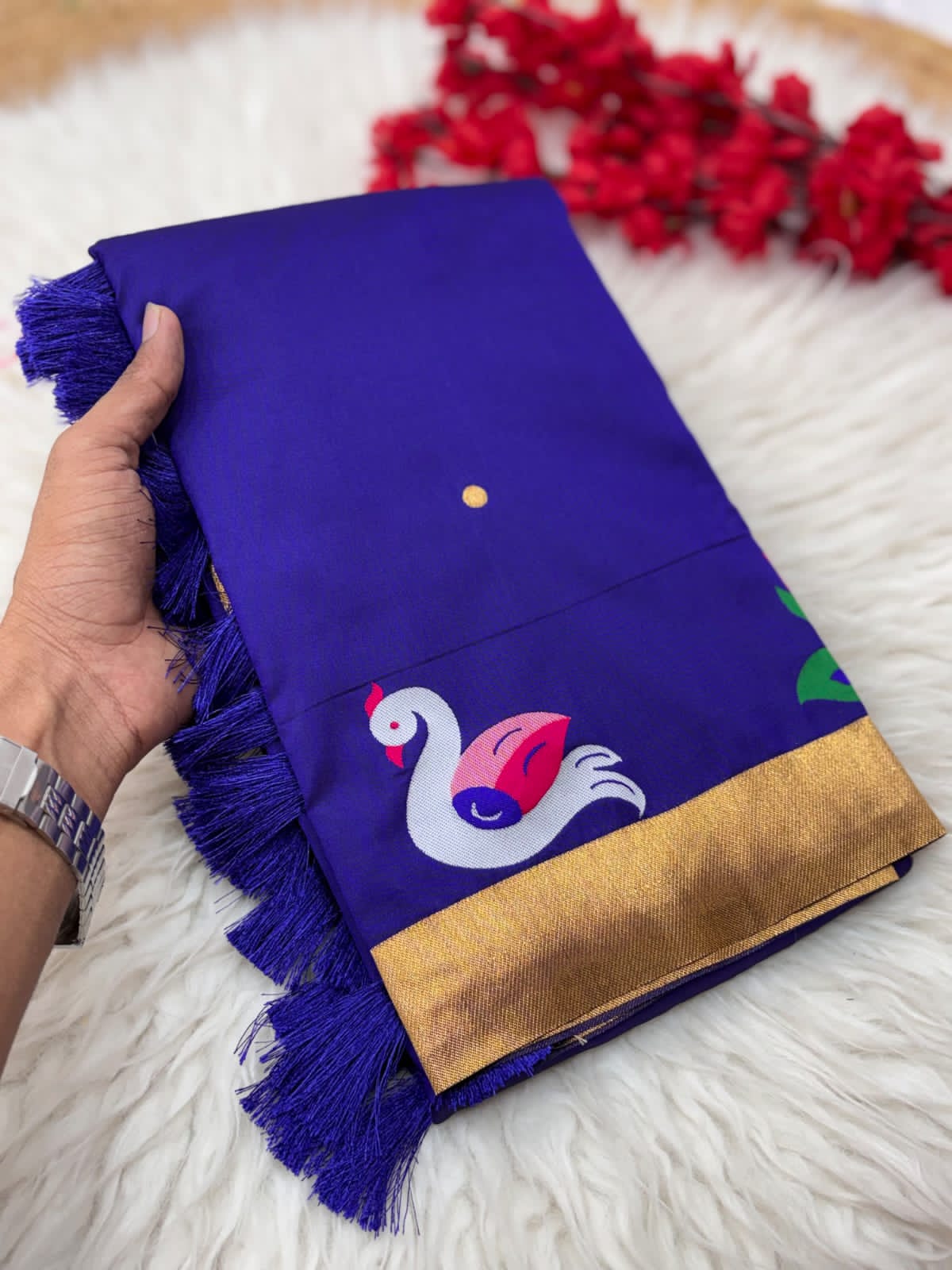 Royal blue saree with gold border and swan design held by a hand on a surface with  flowers in the background.