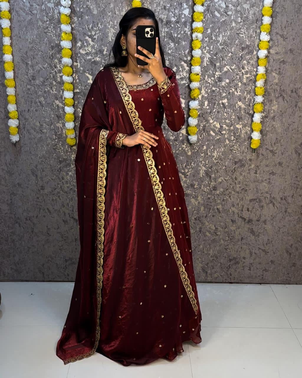 Person wearing a maroon traditional outfit with gold embroidery against a decorative wall.
