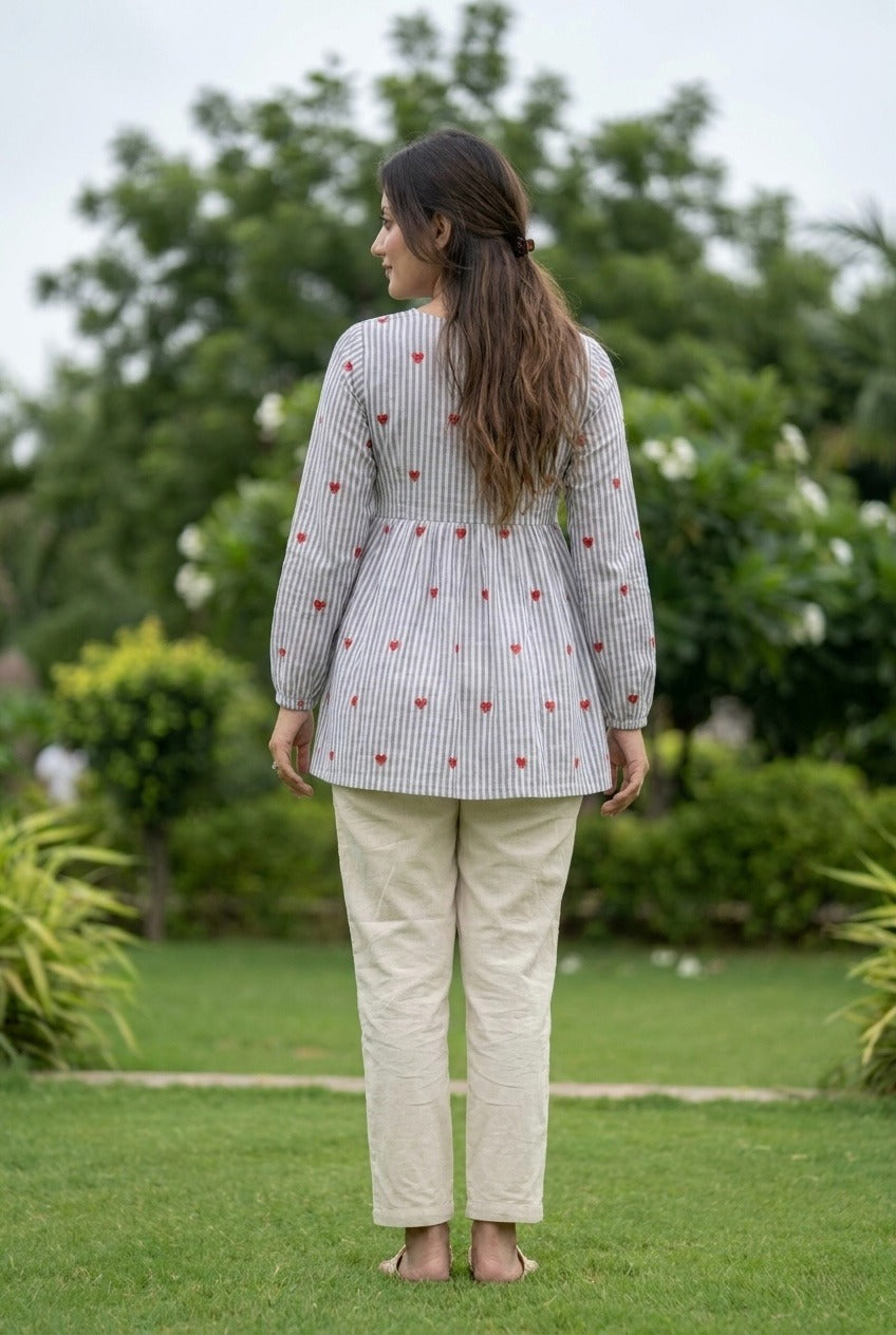 Woman standing outdoors wearing a gray patterned top and beige pants with greenery in the background