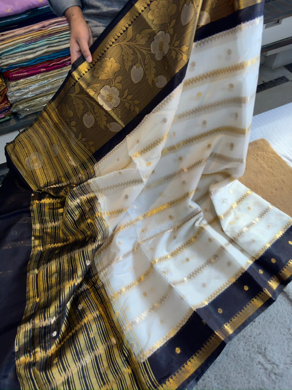 Saree with black and gold border held by a person, with a blurr background of more sarees.