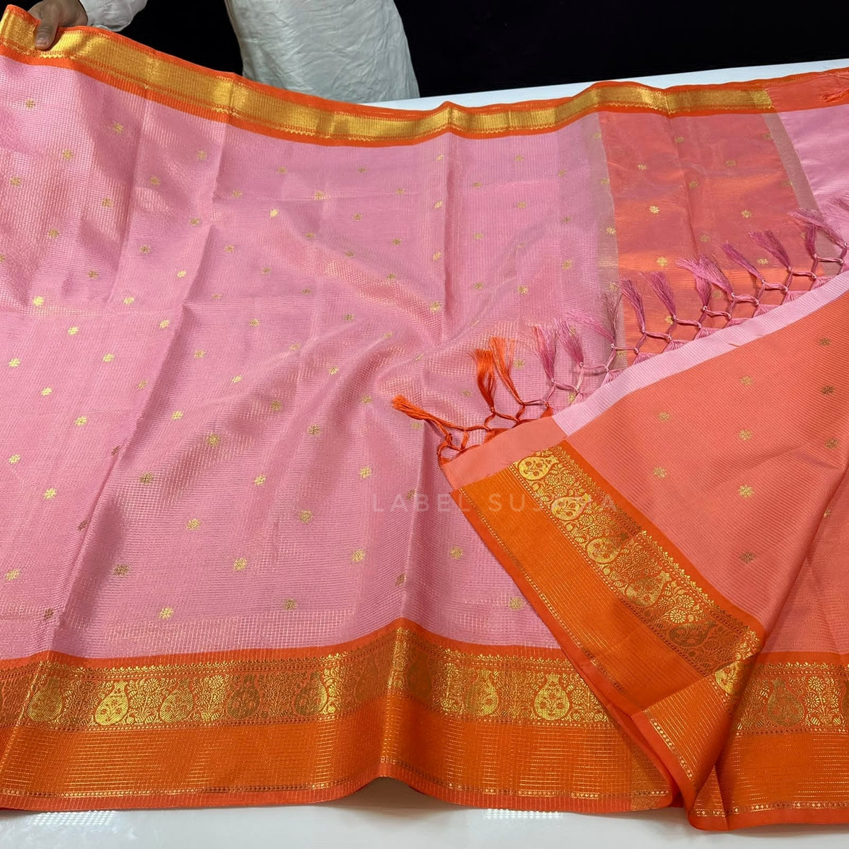 A pink and orange silk saree with gold zari motifs and borders, held out to display its pattern and texture.