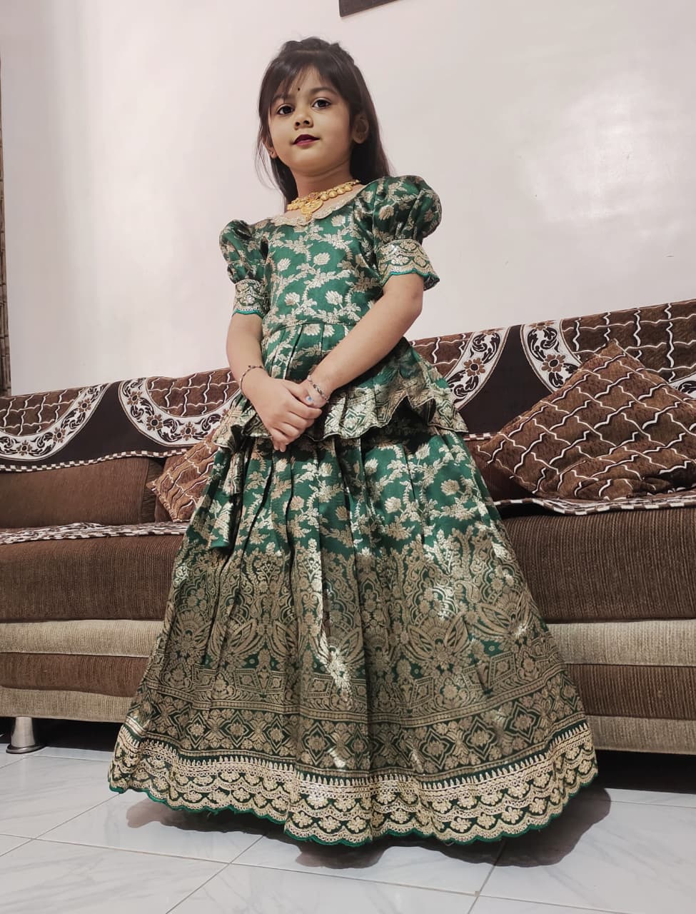 Young girl in a bottle green patterned dress standing in a room with a brown sofa.