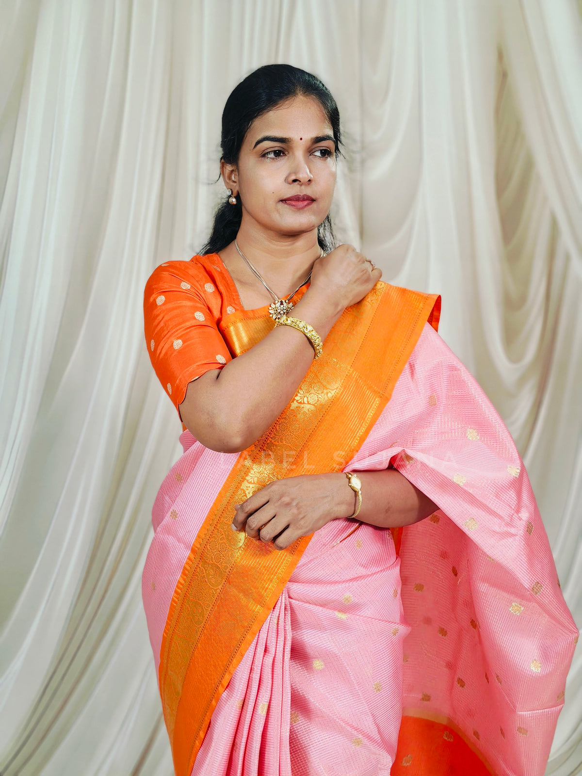 A woman wearing a pink and orange saree with gold motifs, standing in front of a draped cream fabric backdrop.