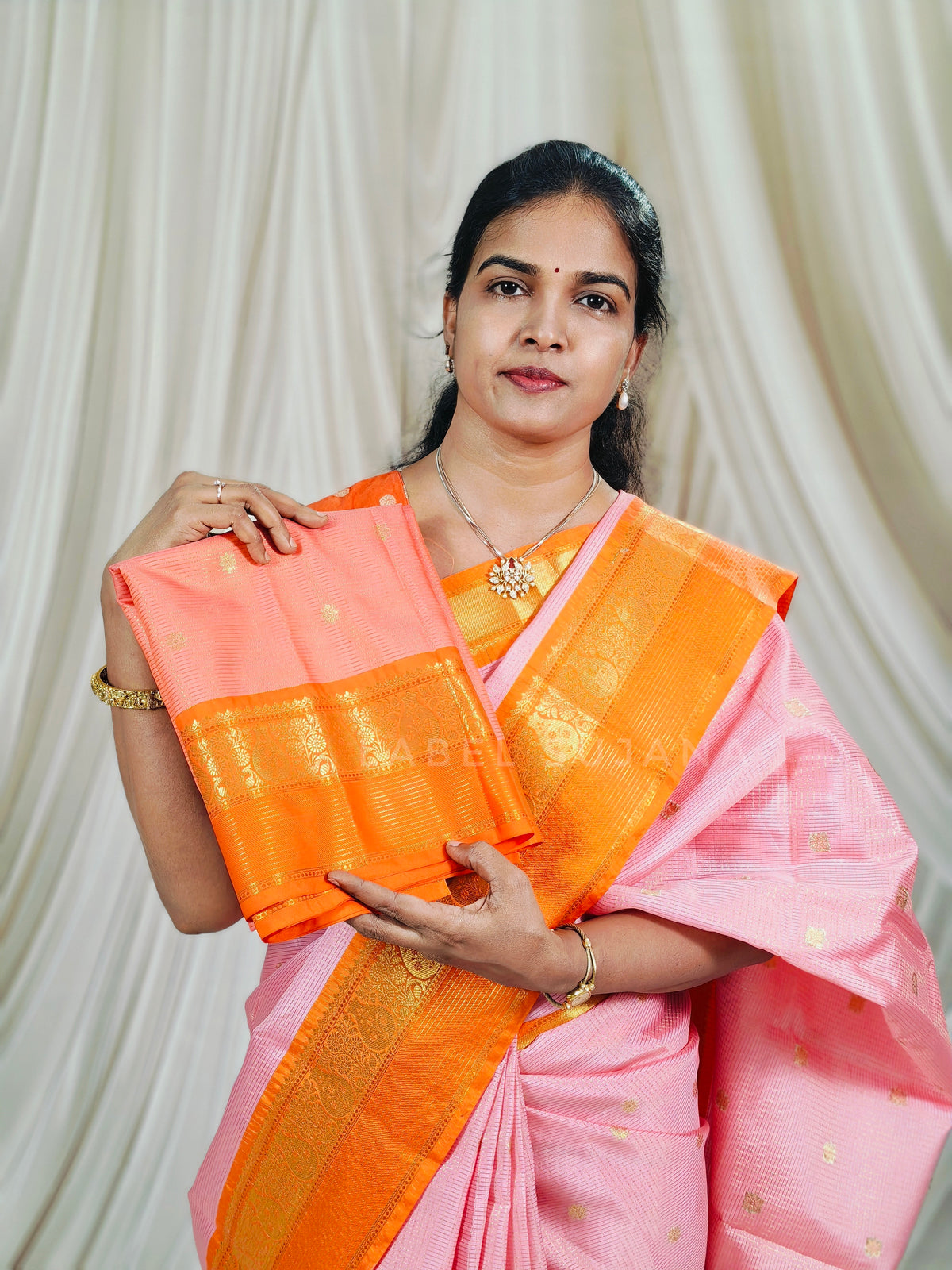 A woman wearing a pink and orange saree with gold zari embroidery, holding up a folded section of the fabric to display its pattern and texture against a draped cream backdrop.