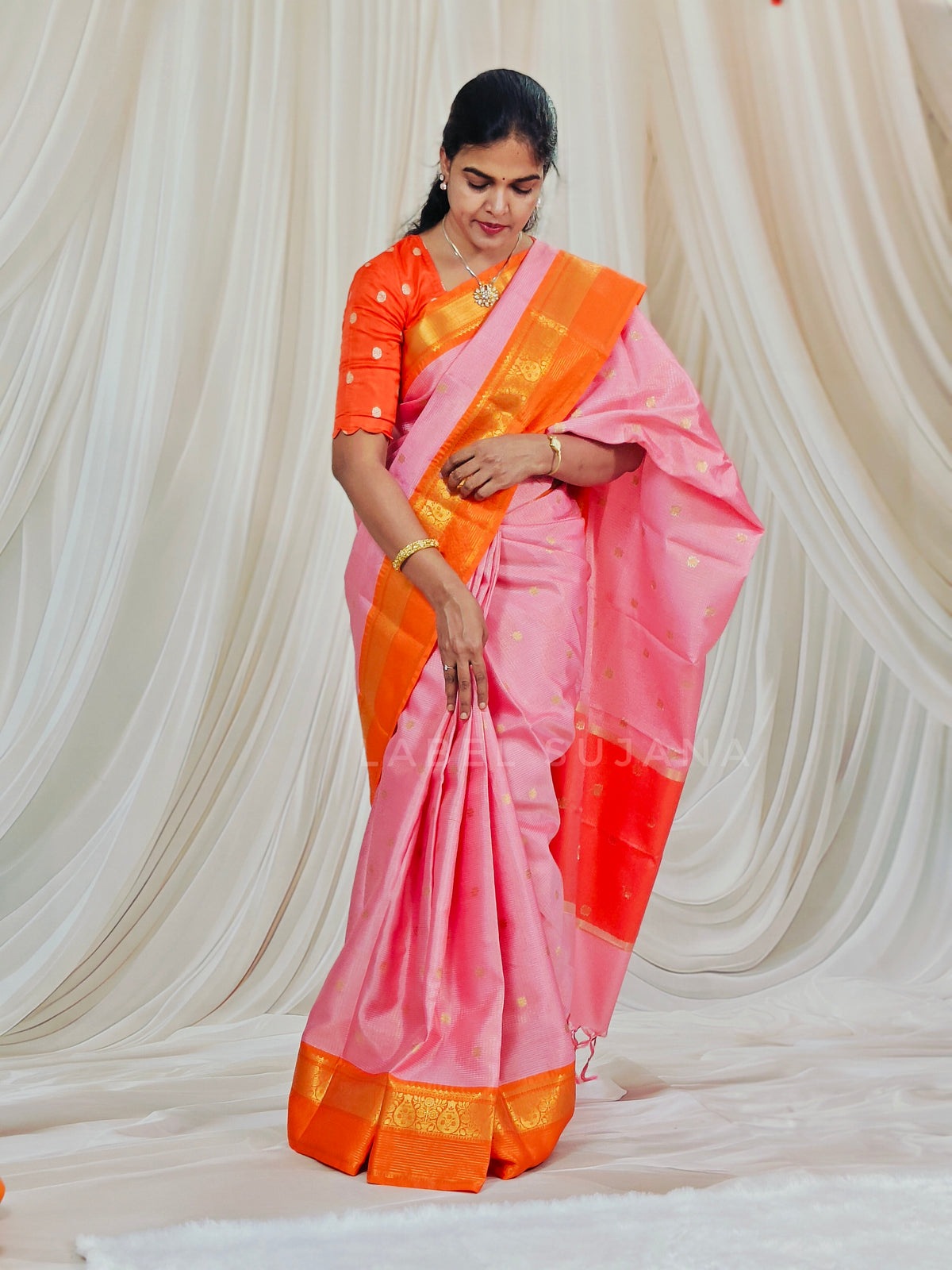 A woman wearing a pink and orange saree with gold zari detailing, standing in front of a draped cream fabric backdrop.