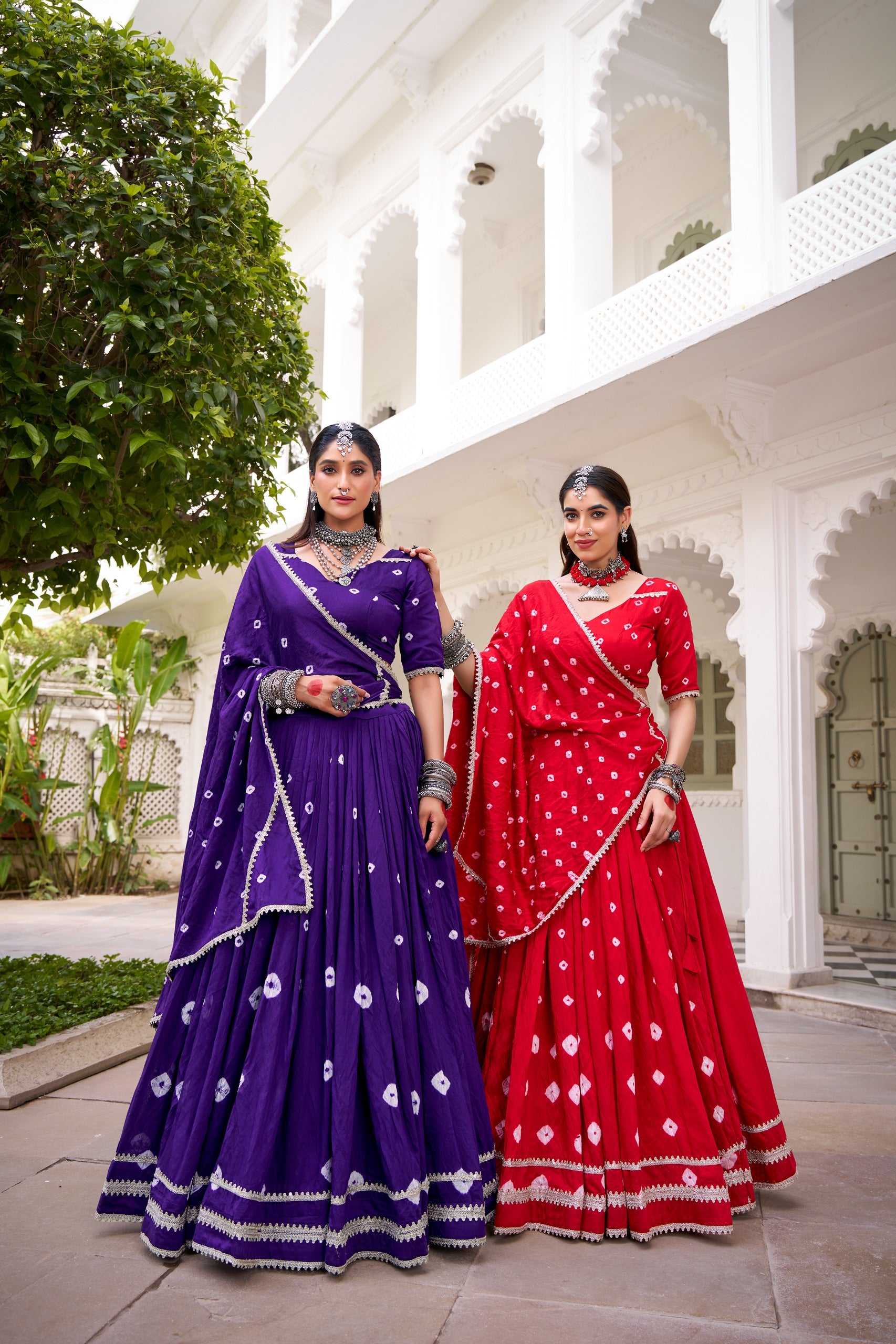 Two women in traditional purple and red lehengas standing in front of a white architectural building.
