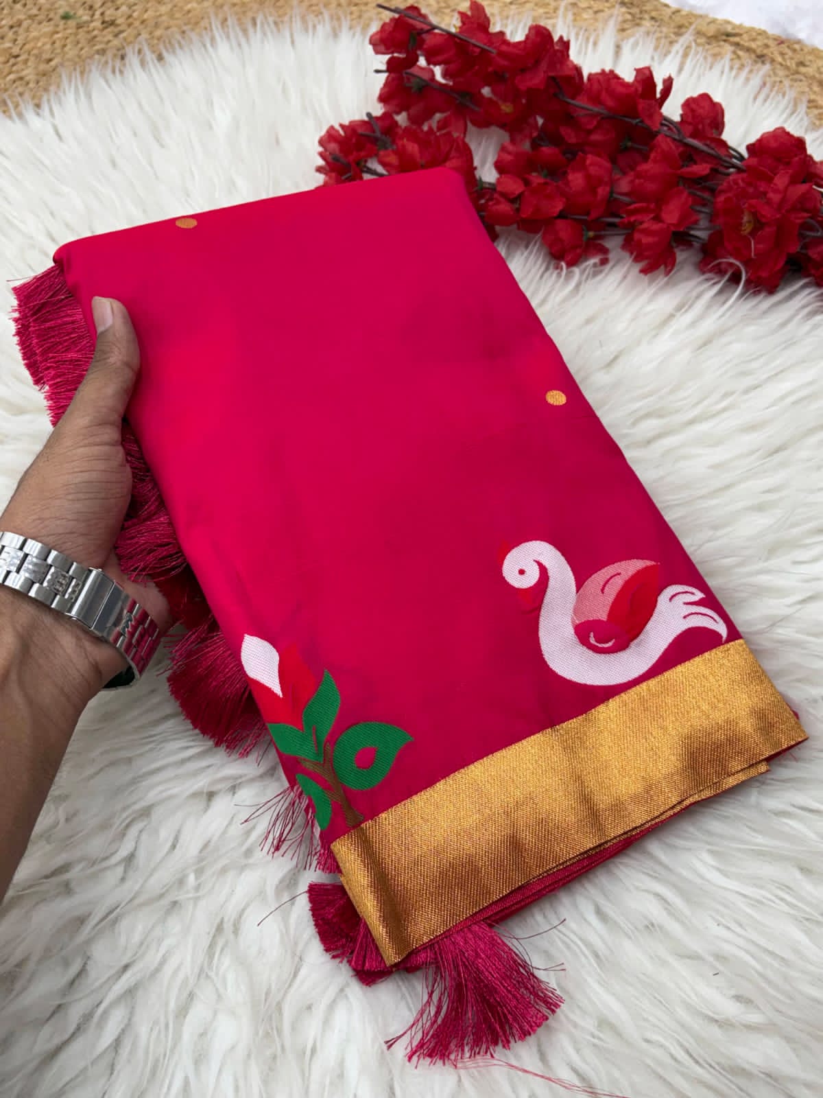 Pink saree with gold border and decorative elements held by a hand on a surface with flowers.