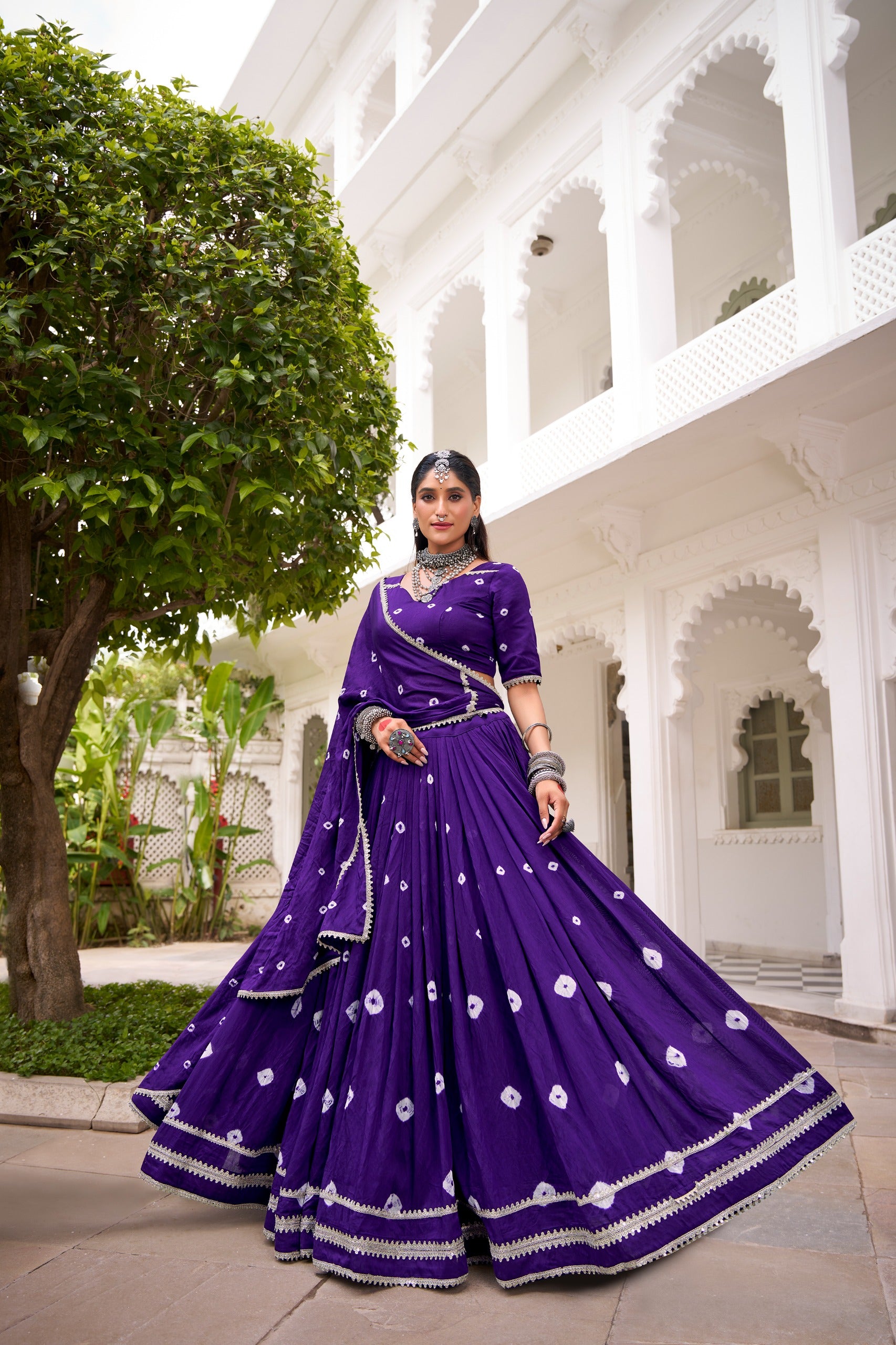 Woman in a purple traditional outfit standing in front of a white building with greenery.