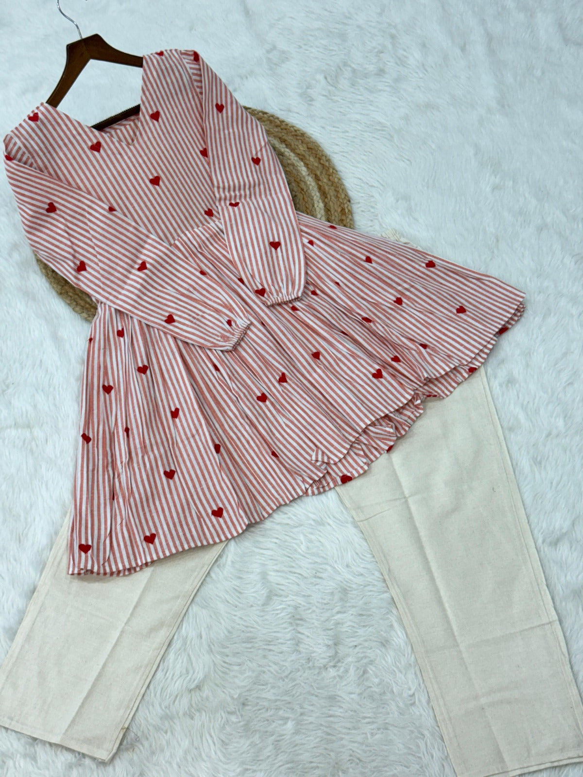 Baby pink and white striped shirt with heart patterns and beige pants on a textured surface