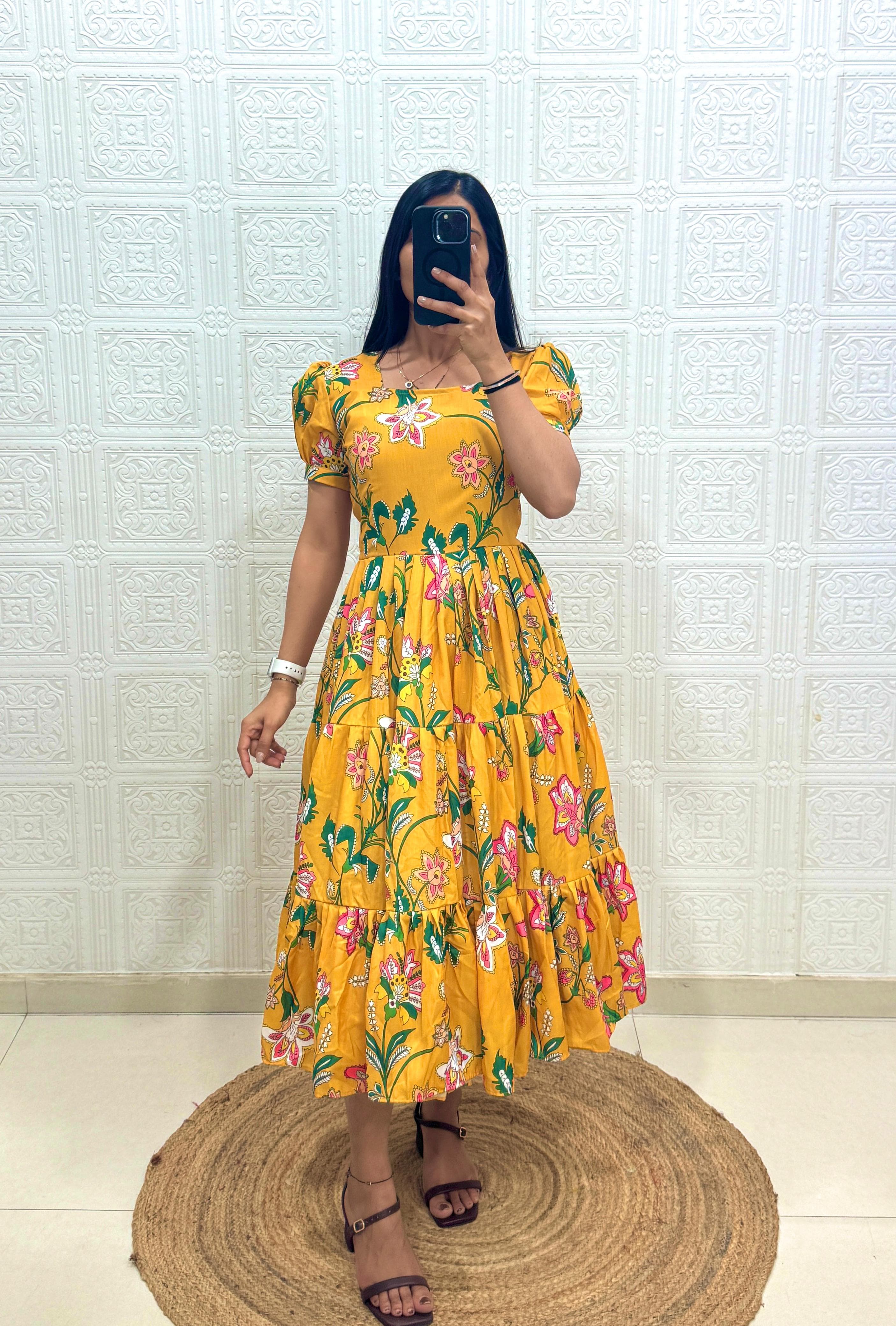 Woman wearing a yellow floral dress taking a mirror selfie against a white tiled wall.