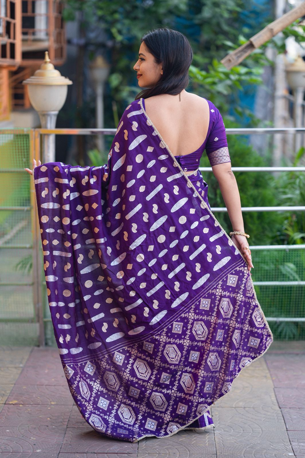 Woman wearing a purple saree with a geometric pattern, standing outdoors.