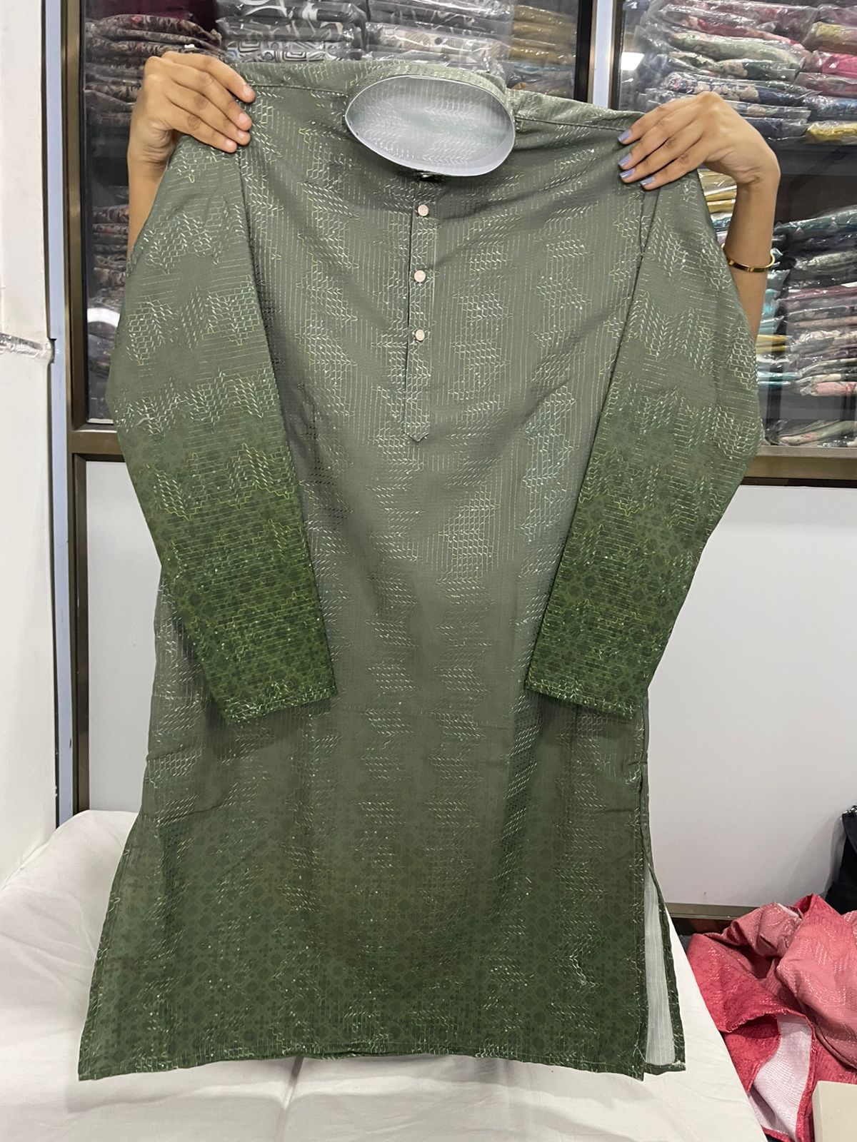 Olive Green kurta being held up against a blurred background of clothing racks.