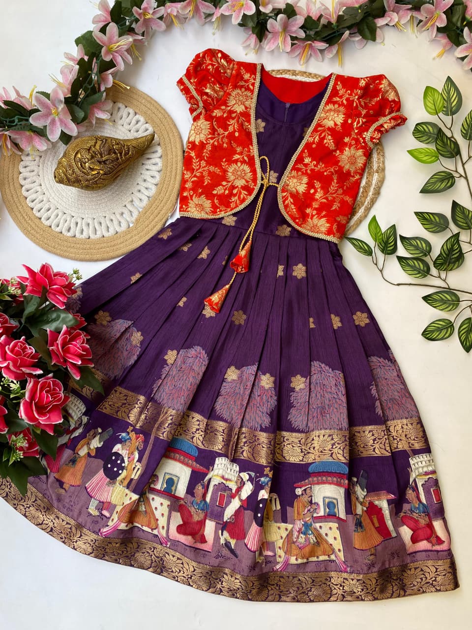 Traditional outfit with red blouse and purple skirt featuring artistic designs, surrounded by flowers and leaves.