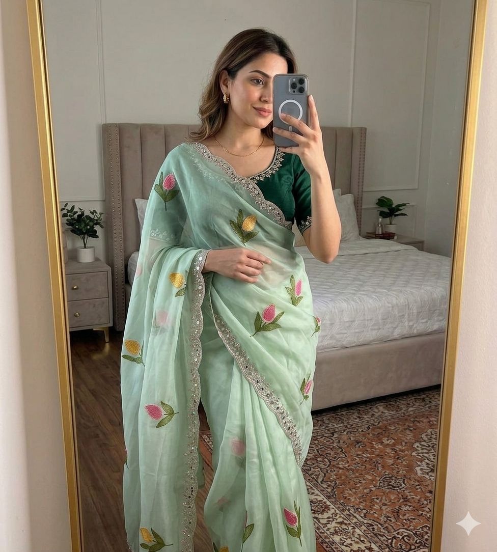 Woman taking a mirror selfie wearing a light green saree with floral patterns in a bedroom.