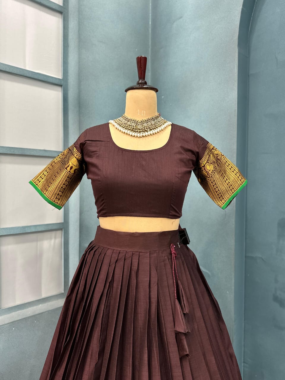 Brown traditional outfit with gold and green details on a mannequin against a light blue curtain background.